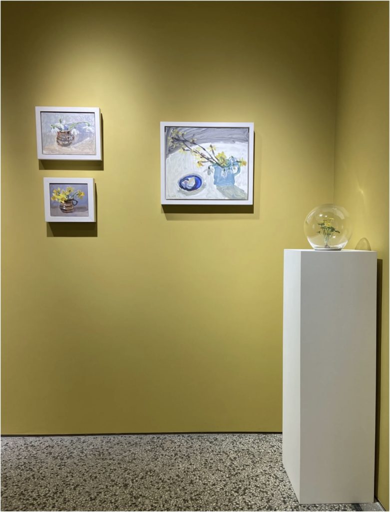Wax flower in a glass orb on tall plinth beside artworks displayed on a yellow wall